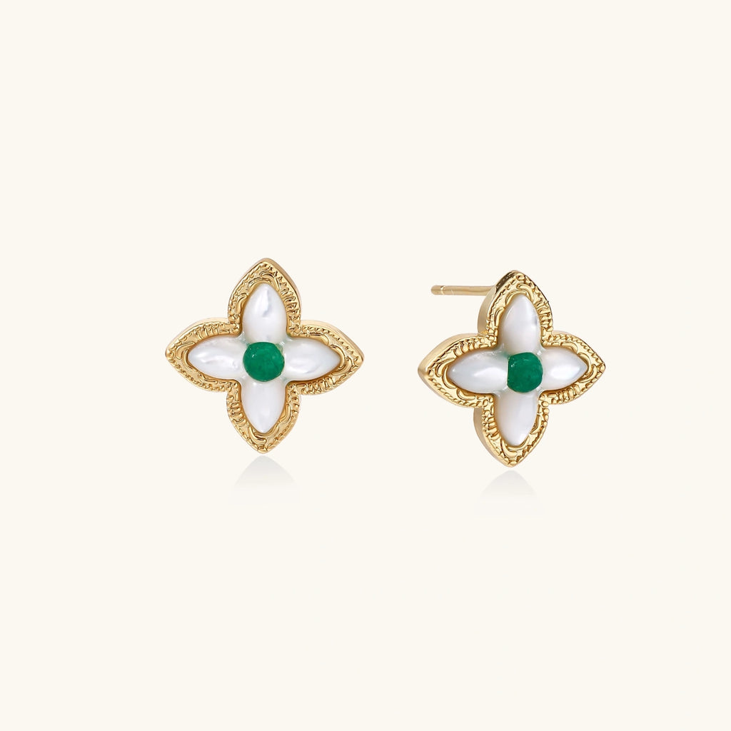 Gold quatrefoil stud earrings with white mother-of-pearl petals and emerald green gemstone center on beige background.