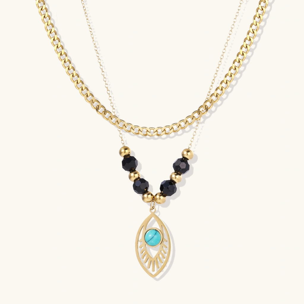 Layered gold necklace with turquoise evil eye pendant and black quartz beads on cream background