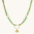 Layered necklace with green stone beads, gold spacers, white accents, and protective eye pendant on cream background.